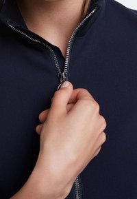 Marineblaue Zip-Jacke mit silbernem Reißverschluss, enganliegendem Kragen und glatter Textur, mit einer Hand, die den Reißverschlussgriff hält.