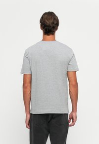 Mann med kort brunt hår iført en enkel lysegrå t-skjorte med korte ermer og mørke jeans, vist fra ryggen mot en hvit bakgrunn.