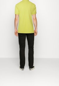 Polo jaune à manches courtes avec une texture lisse, associé à un pantalon noir et des chaussures foncées, vu de dos.