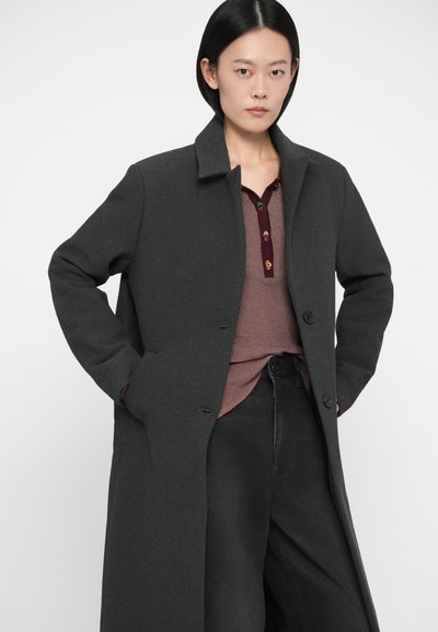 Frau mit schwarzen Haaren, die einen langen dunkelgrauen Mantel, ein mauvefarbenes, geripptes Oberteil mit bunten Knöpfen und schwarze Hosen trägt, steht mit den Händen in den Taschen.