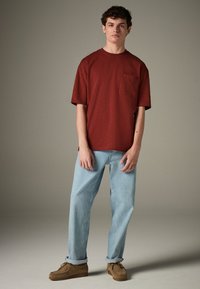 Tricou roșu cu mâneci scurte și buzunar frontal, combinat cu blugi drepti de culoare albastru deschis și încălțăminte maro din piele întoarsă. Fundal simplu.