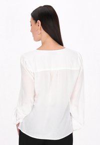 Blusa bianca a maniche lunghe con scollatura rotonda, tessuto morbido, dettaglio arricciato sul retro e maniche leggermente a sbuffo, indossata con pantaloni neri.