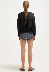 Svart långärmad topp med samlad midja, kombinerad med blekta denimshorts. Modellen har på sig platta svarta sandaler och håret uppsatt.