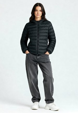 Vrouw staand met handen in de zakken, gekleed in een zwarte pufferjas, losse grijze spijkerbroek en witte sneakers tegen een eenvoudige lichte achtergrond.