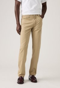 Μπεζ βαμβακερά chino με ίσια γραμμή, διαθέτουν μπροστινές τσέπες και κλείσιμο με κουμπί, συνδυασμένα με σκούρα δερμάτινα παπούτσια.