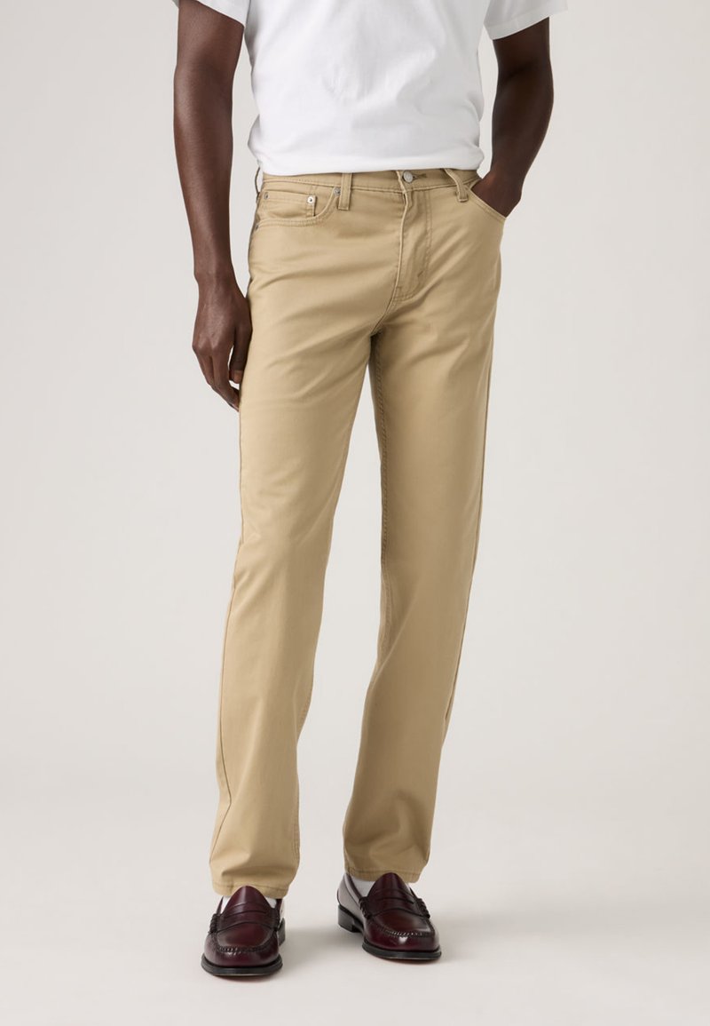 Μπεζ βαμβακερά chino με ίσια γραμμή, διαθέτουν μπροστινές τσέπες και κλείσιμο με κουμπί, συνδυασμένα με σκούρα δερμάτινα παπούτσια.