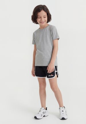 Nasmejan otrok s kratkimi kodrastimi lasmi, oblečen v sivo majico, črne športne hlače z belim robom in bele superge, stoji pred belim ozadjem.