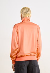 Korallenfarbener Pullover-Jacke mit hohem Kragen, langen Ärmeln, glatter Textur und elastischem Saum, kombiniert mit dunkler Jeans.