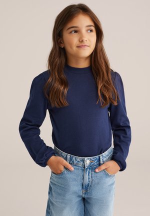 Marineblaues Langarmshirt mit hohem Kragen und strukturiertem Muster, kombiniert mit hellblauen Jeans im High-Waist-Stil und mit vorderen Taschen.