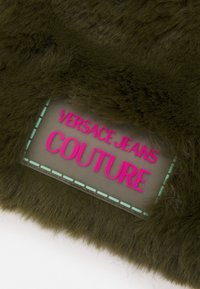 Khaki-färgad konstfårskinn med en framträdande genomskinlig etikett med texten "VERSACE JEANS COUTURE" i fet rosa bokstäver och akvagröna sömnadsdetaljer.