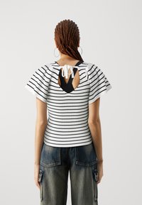 Top de rayas blancas y negras, con un diseño de canalé, mangas cortas con volantes y una abertura en forma de lágrima en la espalda, sujetada con un lazo de cinta blanca.