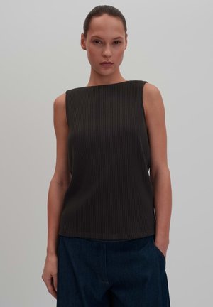 Donna con i capelli raccolti indietro che indossa un top nero a coste senza maniche e pantaloni blu scuro su uno sfondo chiaro uniforme.