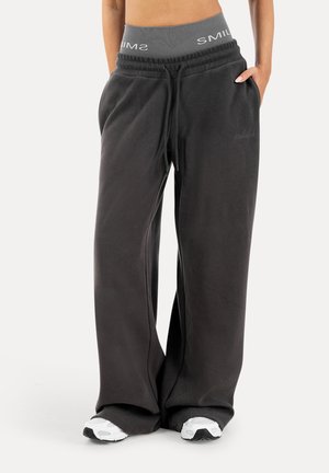 Mujer con pantalones de chándal negros holgados con cintura elástica gris marcada con "SMIL" y zapatillas blancas, de pie con las manos en los bolsillos.