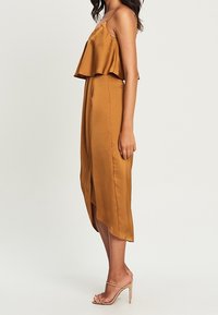 Satijnen bruine jurk met een gelaagde, mouwloze top en een rechte, vloeiende rok. Voorzien van een split aan de voorkant en minimalistische sandalen.