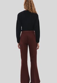 Donna con capelli ricci che indossa una maglietta nera a maniche lunghe e pantaloni marroni a vita alta e a campana, in piedi contro uno sfondo semplice.
