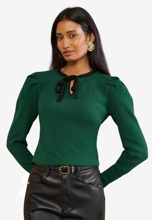 Vrouw met lang zwart haar die een groene trui draagt met zwarte dasdetail en een zwarte leren broek met een gouden gespriem.