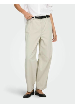 Persona vistiendo pantalones anchos beige con bolsillos delanteros, un cinturón negro, camisa blanca y zapatos planos negros, de pie contra un fondo liso.