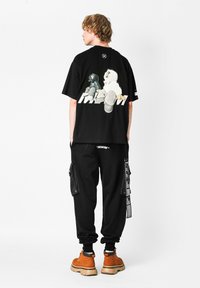 Camiseta gráfica negra con figuras de dibujos animados coloridas en la parte posterior, combinada con pantalones cargo negros y botas de tobillo marrones con cordones.