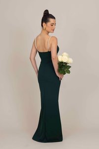 Vestido de noite verde escuro com um design ajustado, alças finas e detalhes em pérolas. A modelo segura um bouquet de rosas brancas.