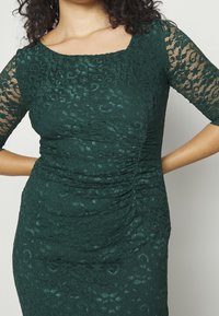 Robe en dentelle vert foncé avec décolleté carré et manches trois-quarts. Présente un tissu texturé avec des motifs floraux et des détails froncés.