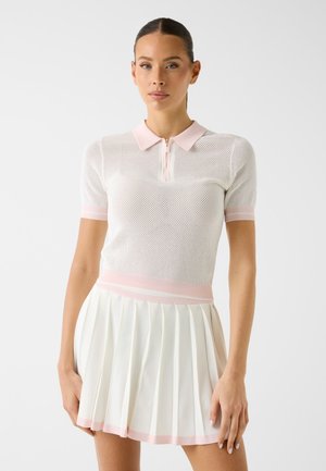 Frau trägt ein weißes Mesh-Poloshirt mit rosa Besatz und einen weißen plissierten Rock mit rosa Rand, steht vor einem einfarbigen Hintergrund.