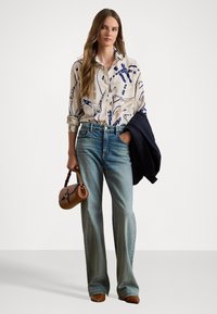 Chemise à boutons avec une base crème et des motifs abstraits bleus, associée à un jean taille haute bleu clair, un sac à main marron et une veste marine.
