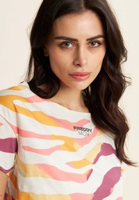 T-shirt a maniche corte color crema con motivi astratti a onde rosa, arancioni e viola; presenta il logo nero "FREDDY MOV" sulla parte anteriore.