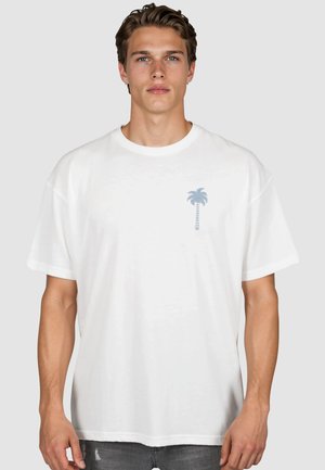 Junger Mann mit hellbraunen Haaren trägt ein locker sitzendes weißes T-Shirt mit einer kleinen blauen Palmen-Grafik auf der linken Brust vor einem einfarbigen Hintergrund.