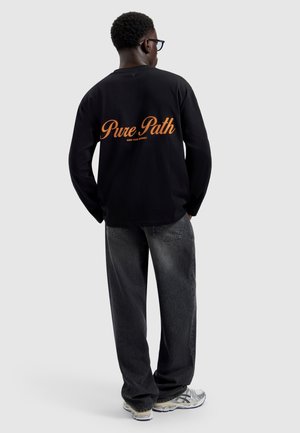 Mann mit kurzen lockigen Haaren und Brille trägt ein schwarzes Langarmshirt mit orangem Schriftzug „Pure Path“, dunkle Jeans und Turnschuhe, blickt weg.