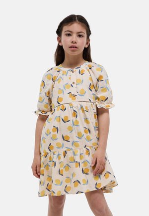 Fille aux cheveux tressés portant une robe crème avec un imprimé citron jaune et bleu, manches bouffantes et boutons sur la poitrine sur un fond uni.