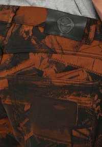 Jeans mit einem orange-schwarzen abstrakten Muster, einem Lederlabel am Bund und einer figurbetonten Passform. Die Textur wirkt robust.