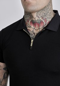Polo negro con cuello con cremallera, que presenta una cremallera dorada y una textura suave. Incluye tatuajes visibles en la zona del cuello.