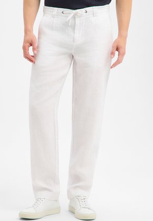 Mann trägt weiße Leinenhose mit Kordelzug in der Taille und weiße Sneaker und steht vor einem einfarbigen Hintergrund.