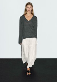 Grauer gestrickter Pullover mit V-Ausschnitt, kombiniert mit einem langen, weißen Faltenrock mit Rauten-Muster. Schwarze Slide-Sandalen runden das Outfit ab.