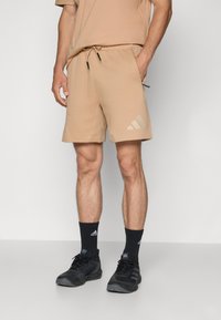 Beige athletic shorts with an elastic waistband and drawstring, featuring a side slit and a subtle three-stripe logo in a lighter shade.