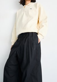 Krämfärgad långärmad polo med två knappar och svart brodyr, tillsammans med löst sittande svarta byxor med en diskret logotyp.