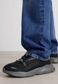 Μαύρα αθλητικά παπούτσια με γκρίζες λεπτομέρειες, που διαθέτουν υφή στην επιφάνεια και στρογγυλή σόλα, φορεμένα με μπλε τζιν παντελόνι.