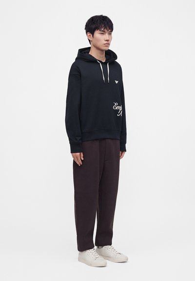 Jeune homme debout portant un sweat à capuche noir avec des cordons blancs, un pantalon ample foncé et des baskets blanches sur un fond clair uni.