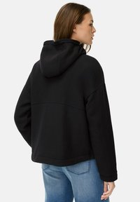 Zwarte hoodie van zacht materiaal, met een relaxte pasvorm, steekdetails op de rug en ribgebreide boorden. Gecombineerd met lichtblauwe jeans.