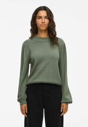 Femme aux longs cheveux bouclés, portant un pull vert à manches longues et un pantalon noir, debout sur un fond blanc uni.