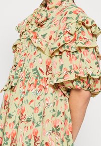 Blumenmusterkleid mit rosa und roten Blumen, Rüschensaum am Kragen und den Ärmeln, getragen von einer Person mit sichtbarem Arm vor einem schlichten Hintergrund.