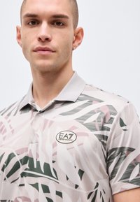 Ung mann iført en mønstret poloskjorte med en grå krage og EA7 Emporio Armani-logo på brystet, stående mot en ensfarget bakgrunn.