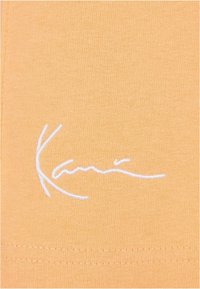 Lyhythihainen, vaalean oranssi t-paita, joka on valmistettu pehmeästä puuvillasta. Tässä paidassa on valkoinen brodeerattu logo vasemmalla alhaalla, ja siinä on sileä pinta.