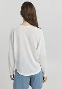 Witte, losvallende trui met een gemêleerd breipatroon, voorzien van een ronde zoom en brede, ontspannen mouwen, in combinatie met blauwe shorts.