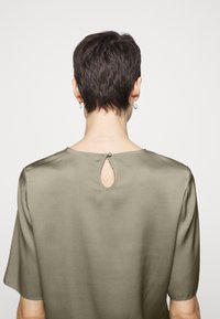 Olivgrünes, kurzärmliges Oberteil aus glattem Stoff, das einen Keyhole-Ausschnitt und einen Knopfverschluss auf der Rückseite hat und einen lockeren Schnitt bietet.