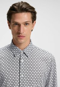 Witte shirt met zwart geometrisch patroon; heeft een button-down kraag en twee knopen aan de voorkant, gemaakt van een gladde stof.