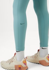 Turkusowe legginsy sportowe z zielonym logo Nike, w połączeniu z beżowymi sneakersami z różowymi i pomarańczowymi akcentami oraz białymi skarpetkami o teksturze.