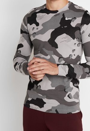 Mann trägt ein graues und schwarzes Camouflage-Langarmshirt mit verschränkten Händen, kombiniert mit dunkelroten Hosen, vor einem schlichten hellen Hintergrund.