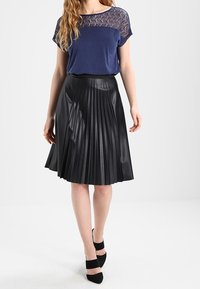 Námořnická modrá blůza s krajkovými detaily a krátkým rukávem, spárovaná s černou skládací sukní vyrobenou z lesklého materiálu. Černé boty s kotníkovými pásky.