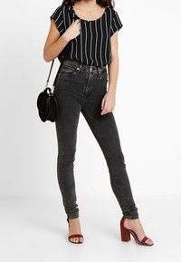 Blusa a strisce bianco e nero con maniche corte, jeans skinny grigio scuro e sandali con tacco blocco bordeaux, abbinati a una borsa a spalla nera.